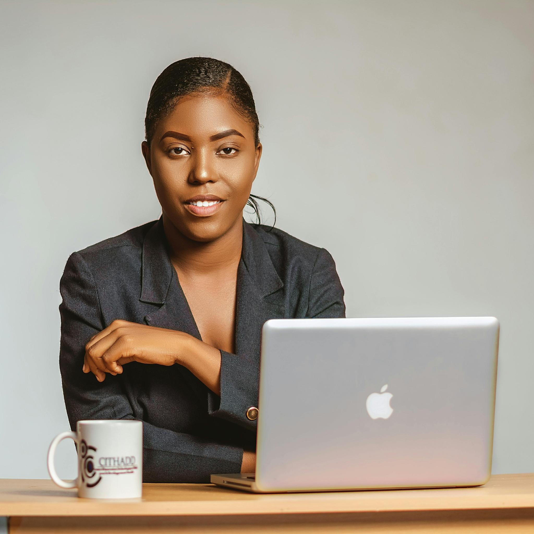 Professional portrait of a woman in a black blazer sitting at a wooden desk with a MacBook laptop and coffee mug. She is smiling confidently at the camera with a neutral grey background behind her. Her hair is neatly styled and she has a polished, business-like appearance.