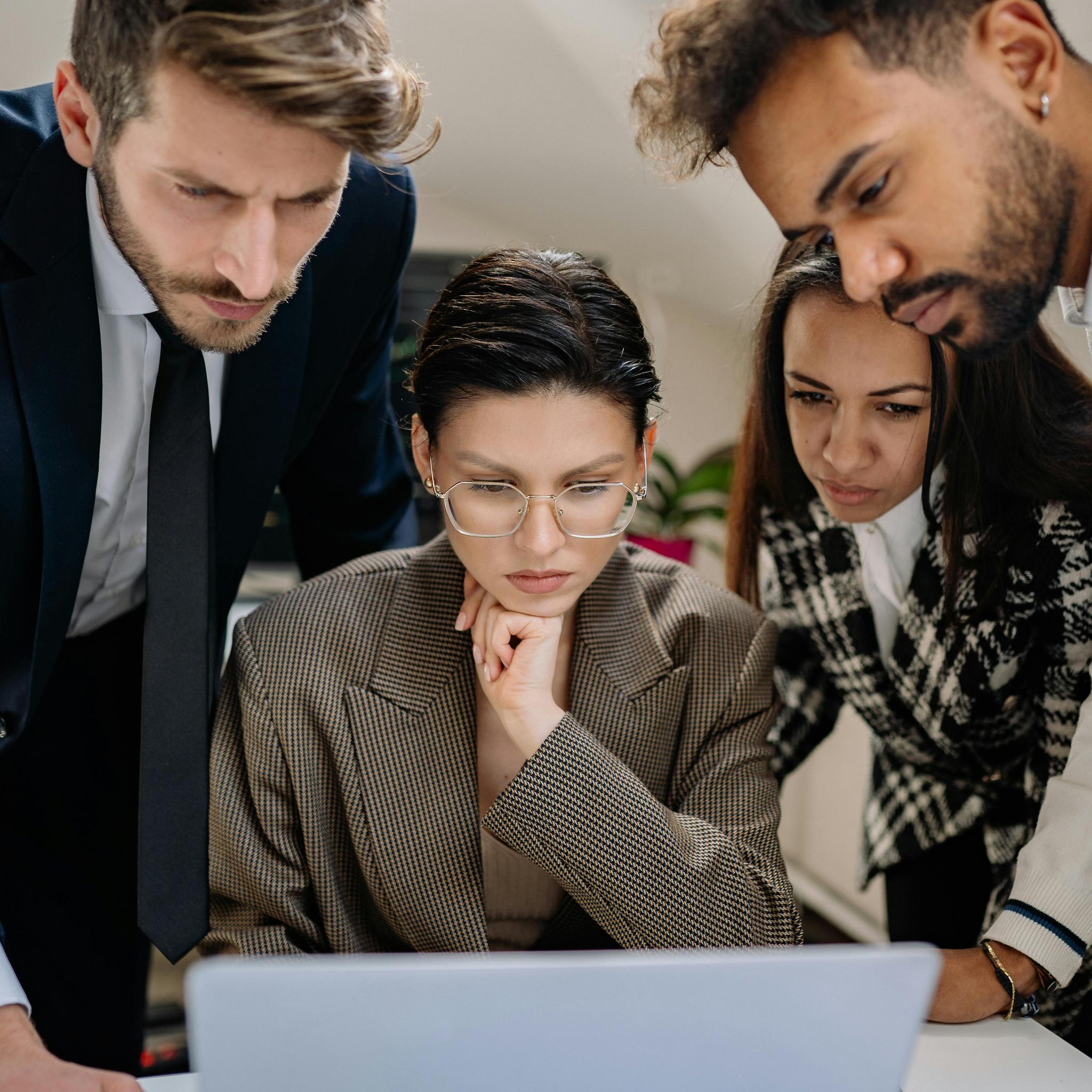 Three business professionals huddle around a laptop, focused intently on the screen. In the center, a person wearing glasses and a checkered blazer rests their chin thoughtfully on their hand. They are flanked by a man in a navy suit and tie on the left, and another colleague in a patterned sweater on the right. The image conveys a collaborative work environment.