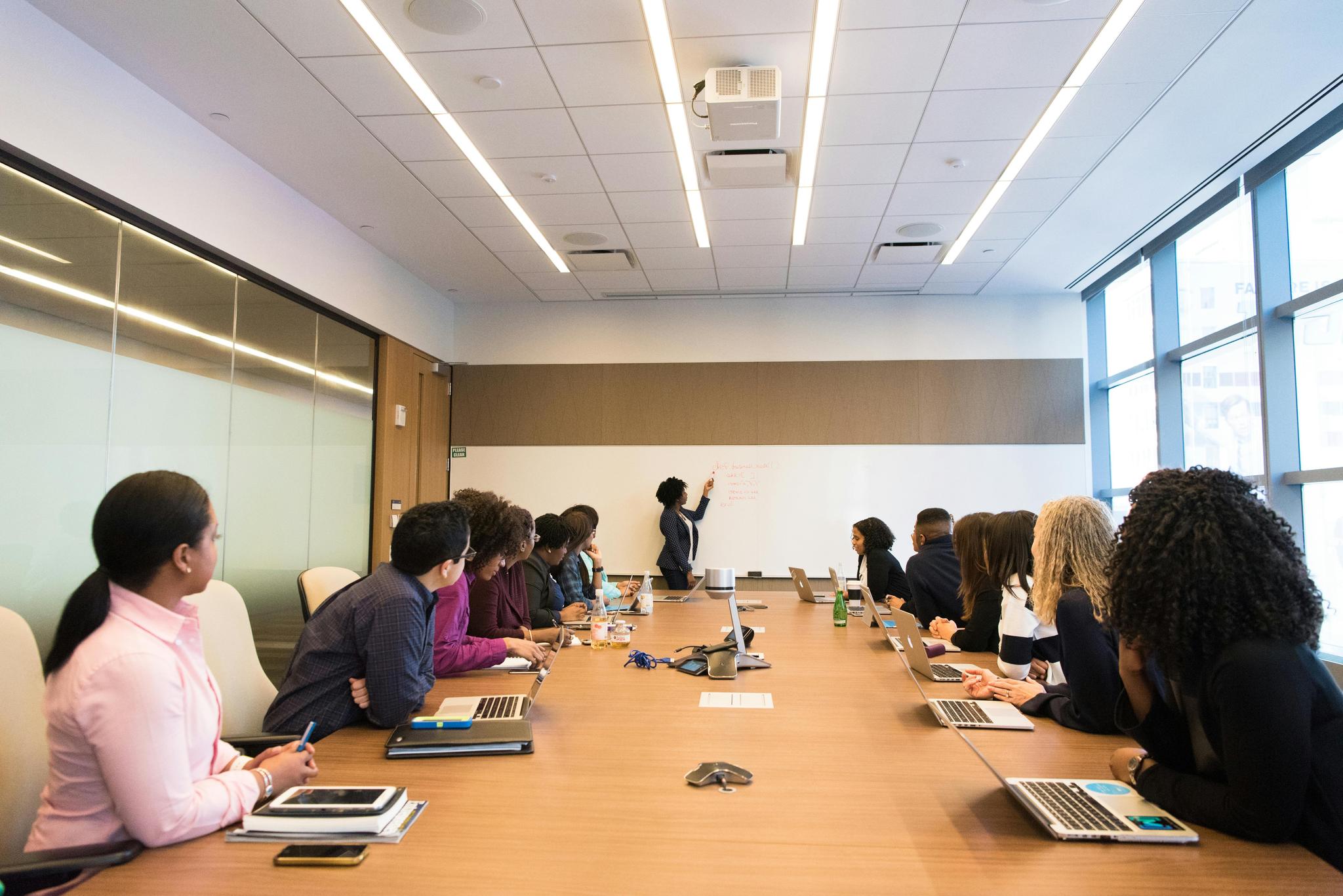 A modern conference room meeting with a presenter standing at a whiteboard while diverse attendees sit around a long wooden table. The attendees have laptops and notebooks, and are seated on both sides of the table. The room features bright fluorescent lighting, floor-to-ceiling windows, and glass partition walls