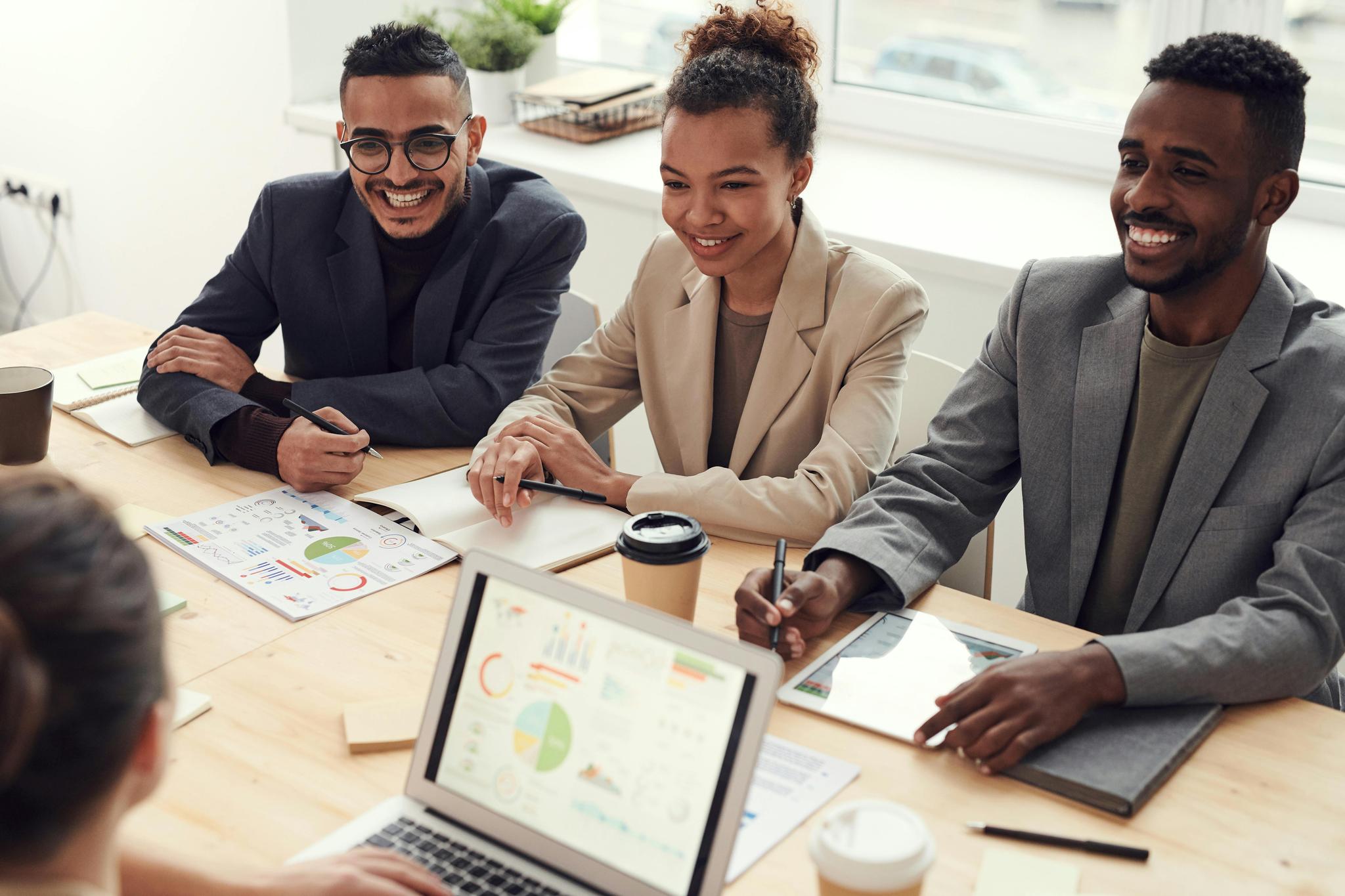 A group of three people are sitting at a table in a meeting room, engaged in a discussion. They have notebooks, pens, and coffee cups in front of them. There are charts and graphs on the table, and a laptop displaying a colorful pie chart and other data.