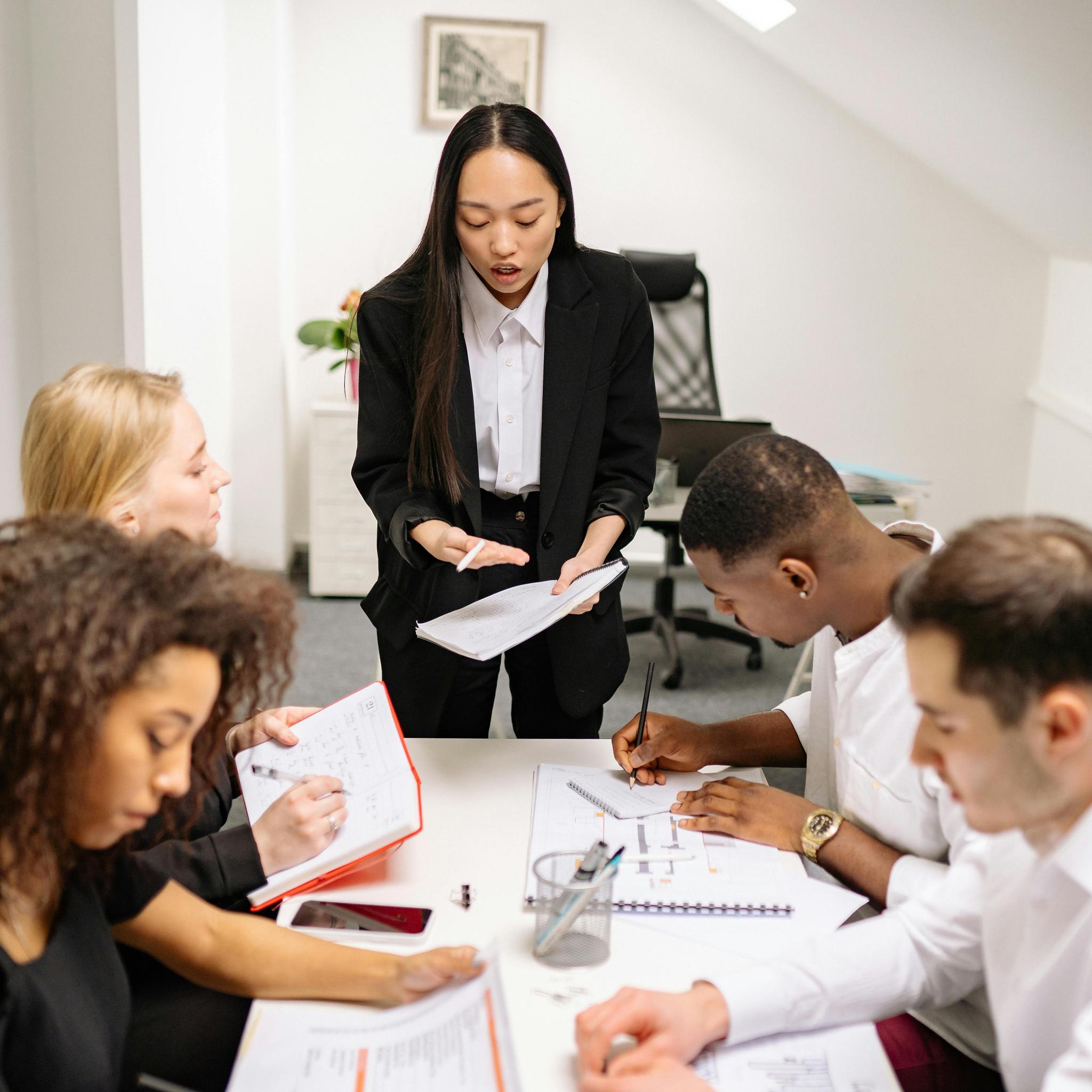 A group of five people are gathered around a table in an office setting. One person, standing and dressed in a black suit, appears to be leading the discussion, holding a notebook and gesturing with a pen. The other four individuals are seated, taking notes and engaging with the material in front of them. Various notebooks, papers, and a smartphone are visible on the table