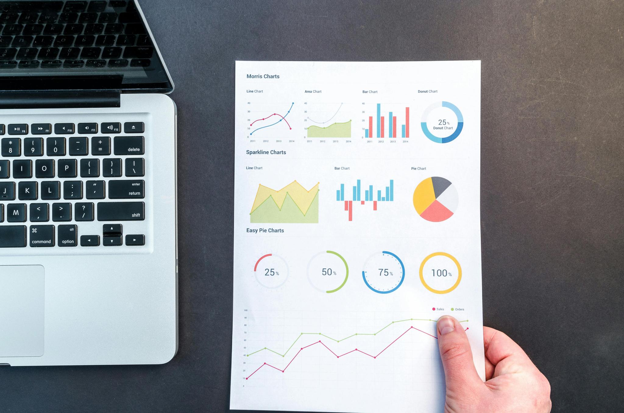 A person holds a sheet of paper with various charts and graphs next to an open laptop. The sheet includes line charts, area charts, bar charts, donut charts, pie charts, and circular progress charts, displaying different types of data visualizations.