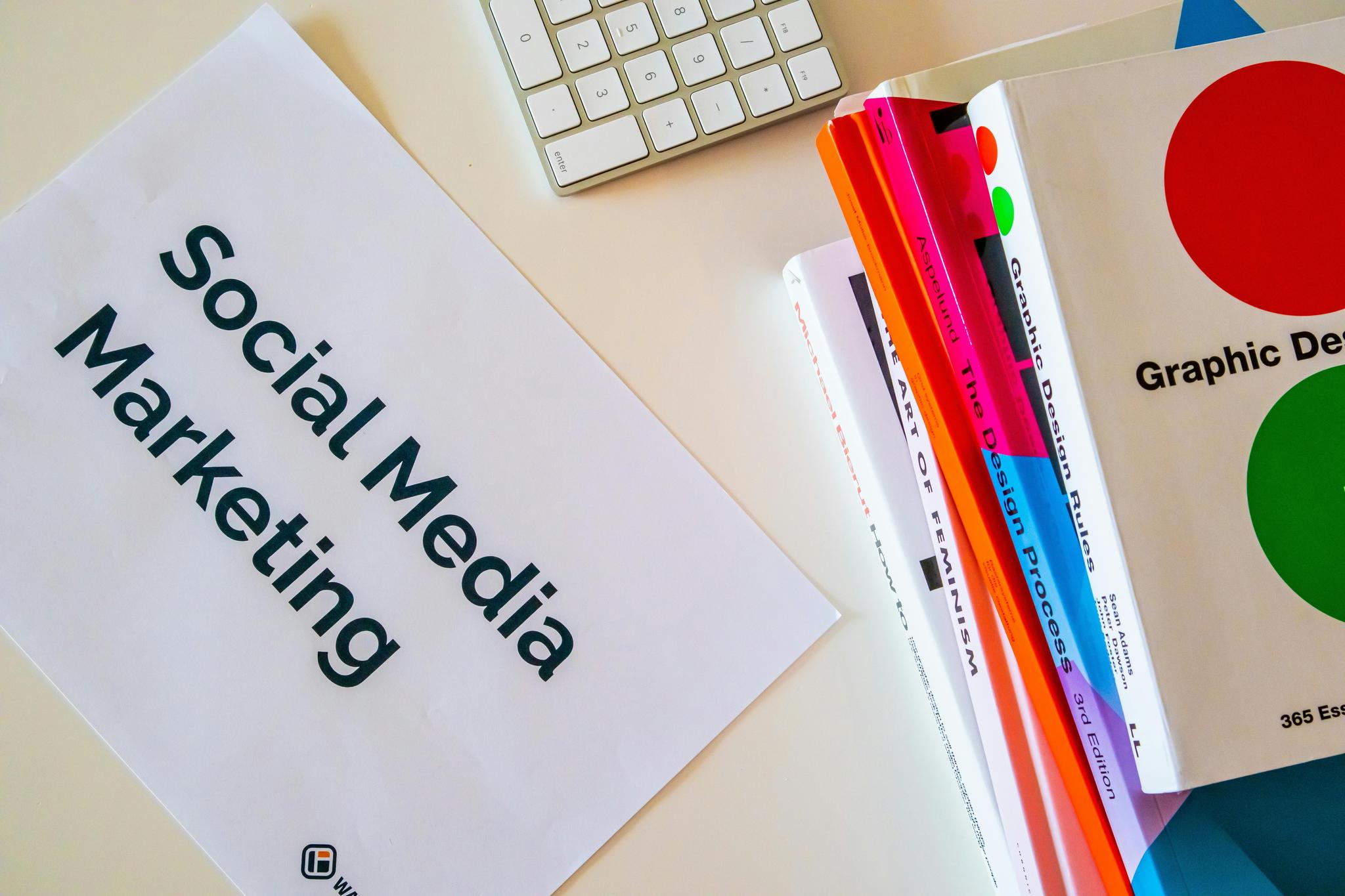 A desk with a sheet of paper labeled 'Social Media Marketing,' a keyboard, and a stack of colorful books. The books include titles on graphic design and feminism.