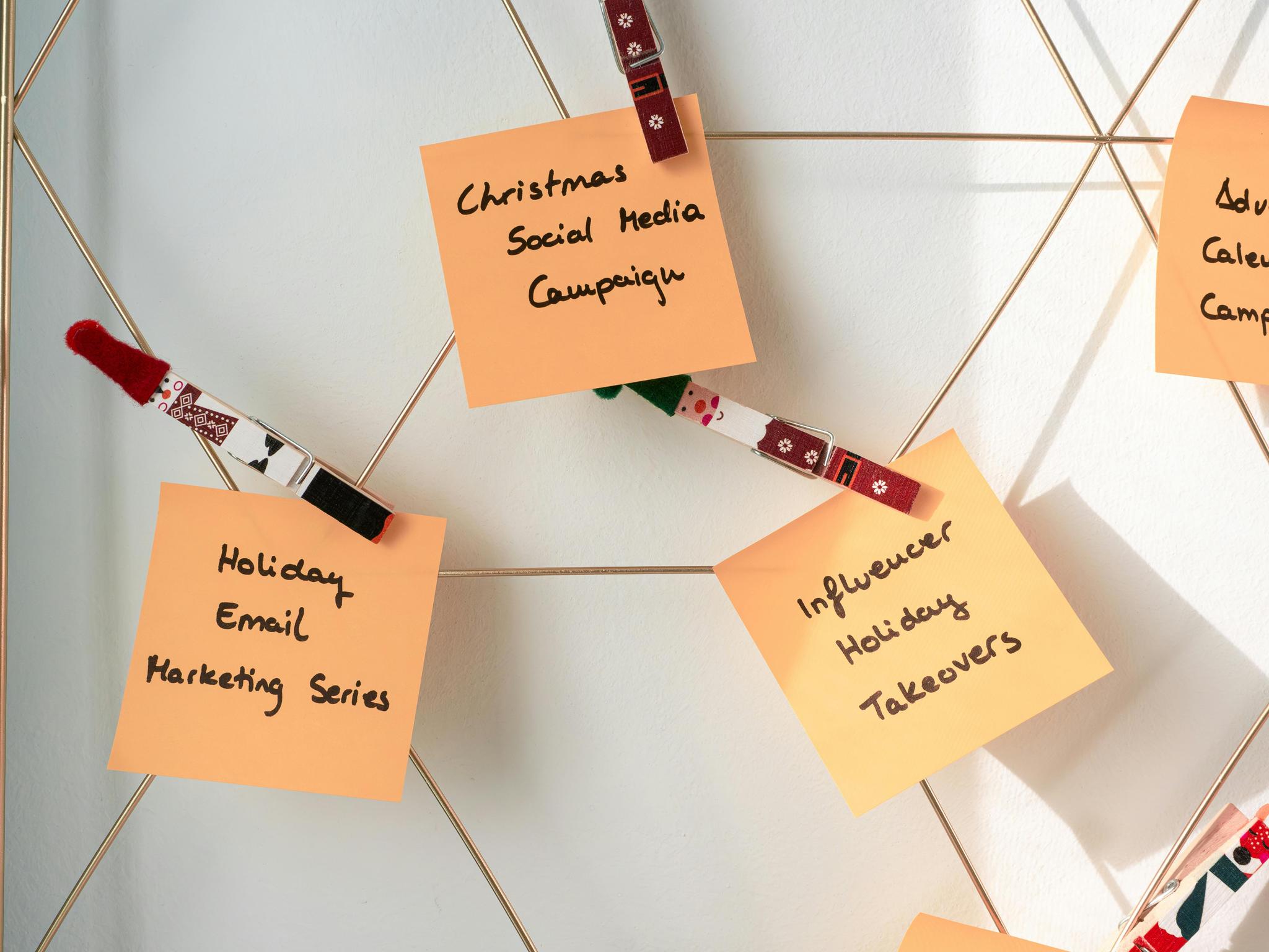 The image shows a wire grid with three sticky notes attached using decorative clothespins. The sticky notes have handwritten text. The first note reads 'Holiday Email Marketing Series,' the second note reads 'Christmas Social Media Campaign,' and the third note reads 'Influencer Holiday Takeovers.' The clothespins have festive designs, including patterns and images of Santa Claus. The notes appear to be part of a planning or brainstorming session for holiday marketing campaigns.