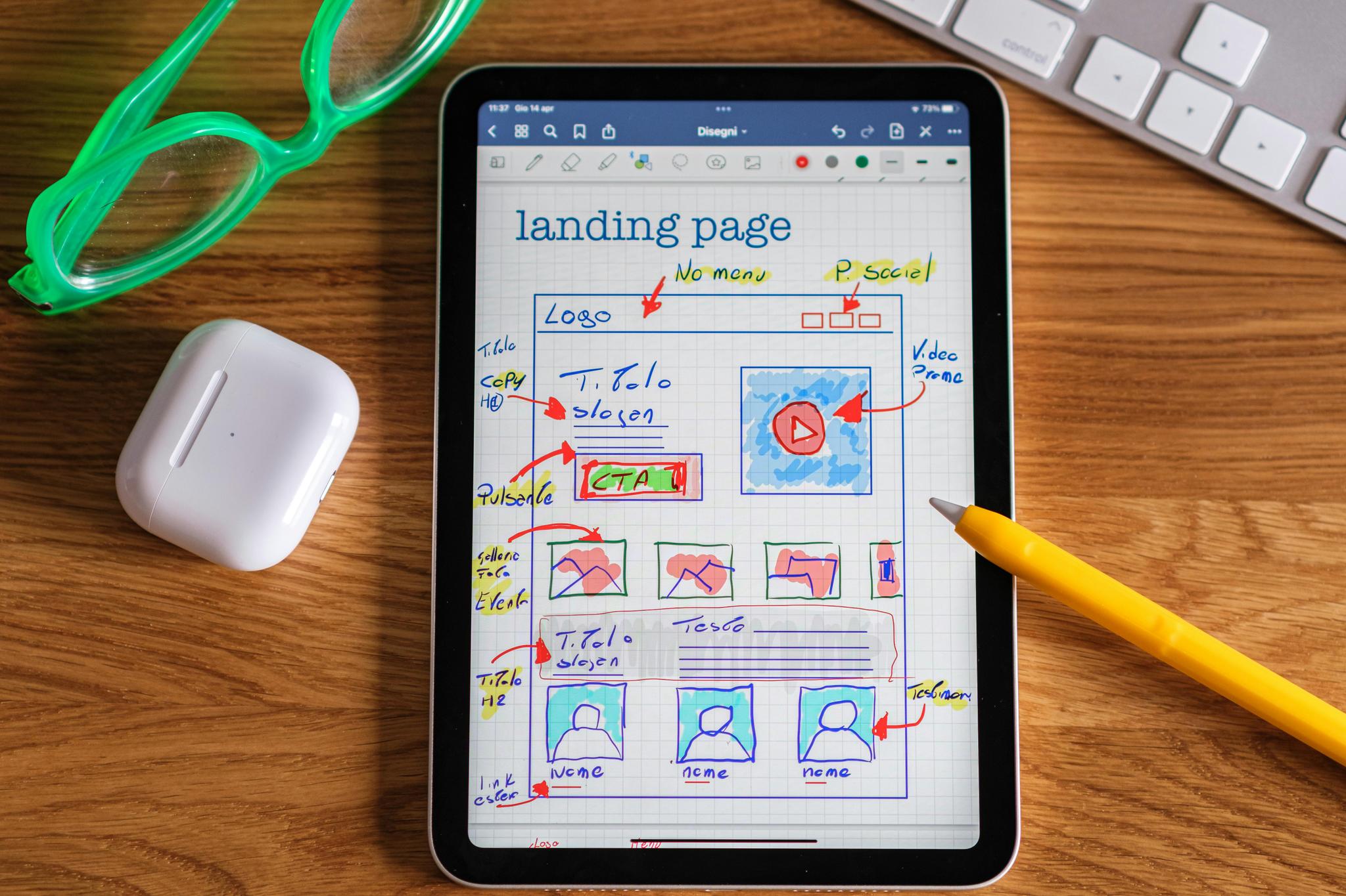 A tablet displaying a hand-drawn sketch of a landing page layout is placed on a wooden desk. The sketch includes various elements such as a logo, navigation menu, social media icons, a video promo section, call-to-action buttons, and placeholders for images and text. Surrounding the tablet are a pair of green glasses, a white wireless earbud case, and a yellow pencil. A keyboard is partially visible in the background.