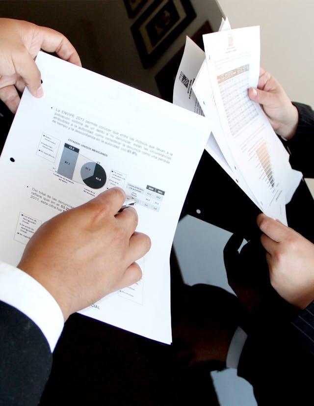 A person holds a sheet of paper with various charts and graphs next to an open laptop. The sheet includes line charts, area charts, bar charts, donut charts, pie charts, and circular progress charts, displaying different types of data visualizations.