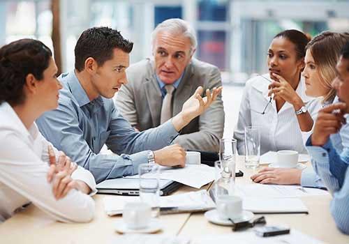 A diverse group of business professionals engaged in an animated discussion around a conference table. A man in a light blue shirt gestures while speaking, with an older man in a grey suit listening intently beside him. Several other colleagues sit across the table, with coffee cups and papers scattered on the surface.